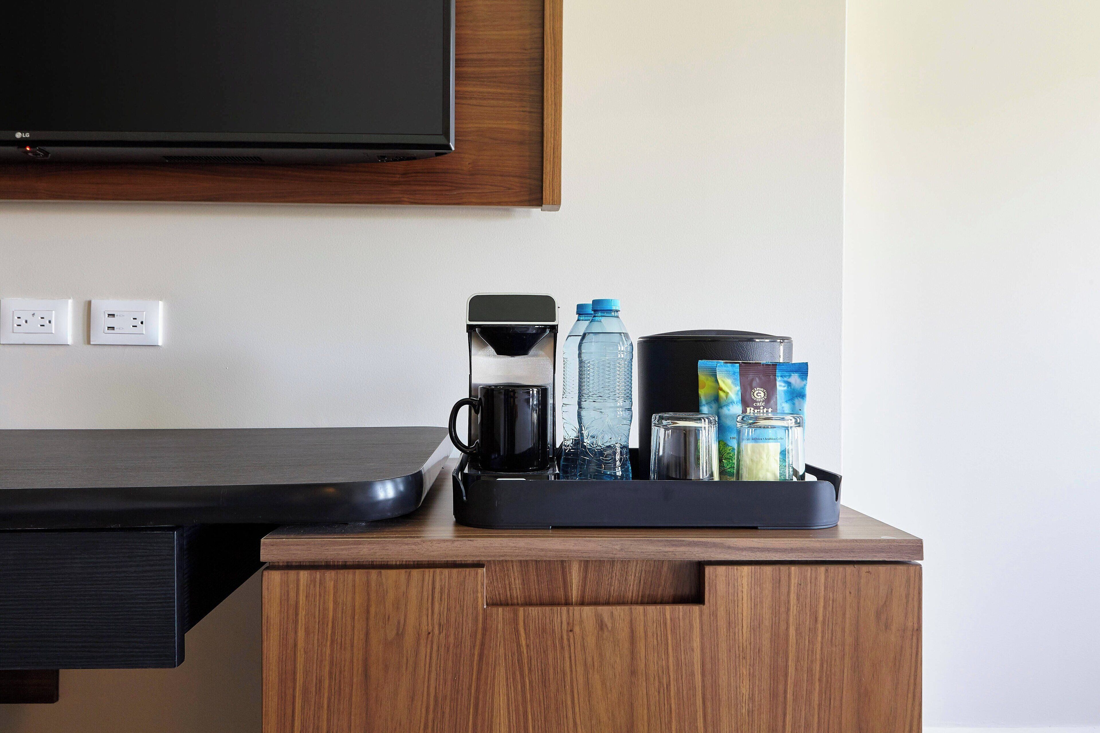 A hotel room with a wooden desk, a flat-screen TV mounted above, and a tray with a coffee maker, water bottles, and a container of snacks.