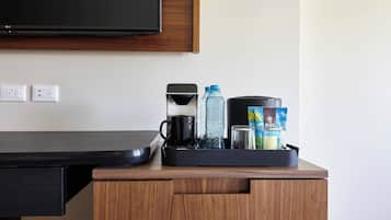 Habitación de hotel con un escritorio de madera, un televisor de pantalla plana montado arriba y una bandeja con cafetera, botellas de agua y un recipiente con aperitivos.