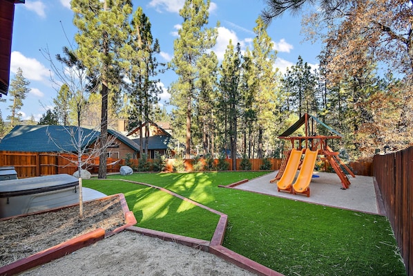 A backyard with a wooden playset, a barbecue grill, and a house with a blue roof.