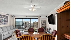 Ein geräumiges Wohnzimmer mit einer grauen Couch, einem Holzesstisch mit vier Stühlen, einem Deckenventilator und einem Balkon mit Blick auf Gebäude.