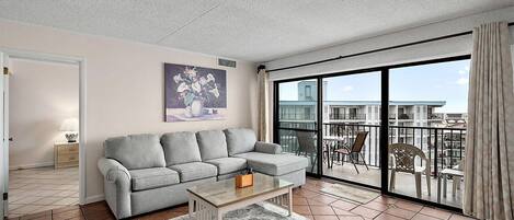 A living room with a grey sofa, a coffee table, and a large sliding glass door leading to a balcony with outdoor furniture.