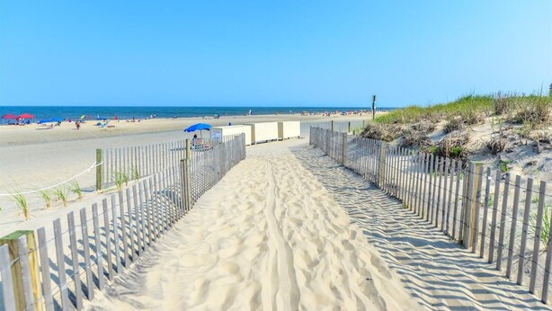 Ein sandiger Küstenweg führt zum Meer, begleitet von einem Holzzaun und Strandsonnenschirmen.