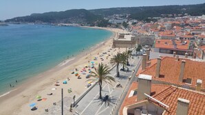 A coastal cityscape with a beach, buildings with red-tiled roofs, and a clear sky. - Música penthouse 360º (Sesimbra)