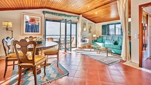 A spacious living area with a wooden ceiling, a dining table with chairs, a sofa, and a large window with a view. - Música penthouse 360º (Sesimbra)