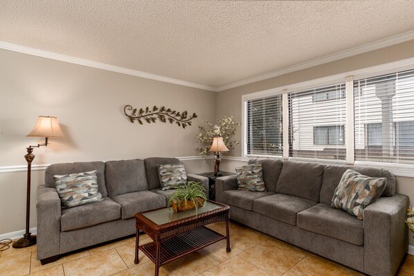 A living room with two grey sofas, a coffee table, a floor lamp, and a decorative wall piece. - Steps to the beach daily rentals pool washer dryer onsite two balconies full kitchen (Myrtle Beach)