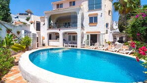 A poolside view of a two-story building with a balcony, outdoor seating, and a clear blue pool.