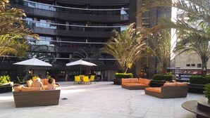 An outdoor seating area with multiple sofas and umbrellas, surrounded by greenery and a modern building in the background.