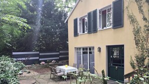 A two-story building with a patio area featuring a table and chairs, a stone pathway, and a fenced garden. - HR Cottage am Honigbach in the center of Coesfeld (Coesfeld)