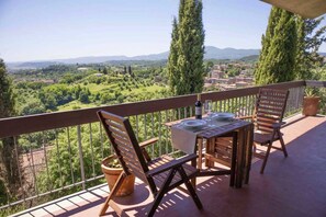 Un balcone con un tavolo apparecchiato per due, affacciato su un paesaggio pittoresco con alberi e edifici.
