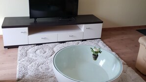 Una sala de estar con un televisor de pantalla plana sobre un soporte negro, un sofá blanco, una mesa de centro de vidrio y una alfombra blanca y mullida.