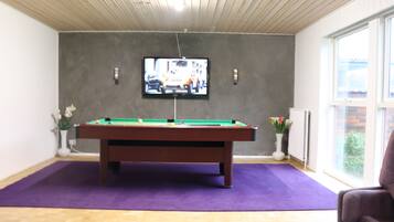 A pool table in a room with a television mounted on the wall, a window, and a chair.