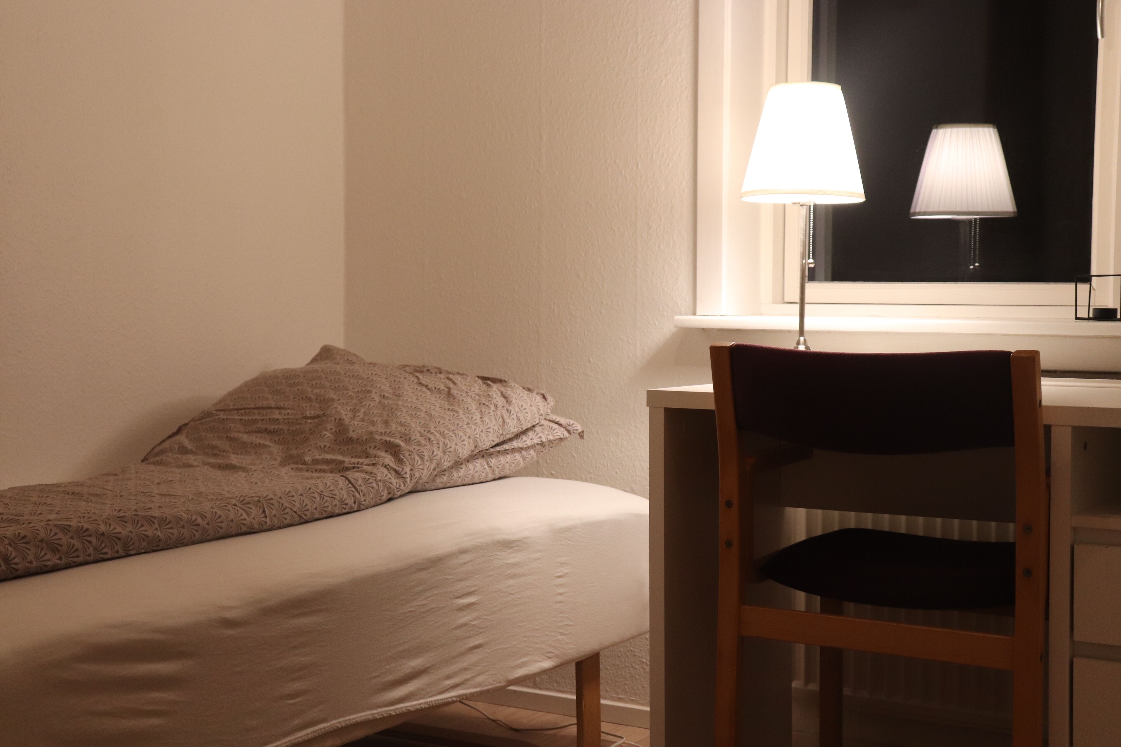 Un letto singolo con coperta a motivi, un comodino con una lampada e una sedia.