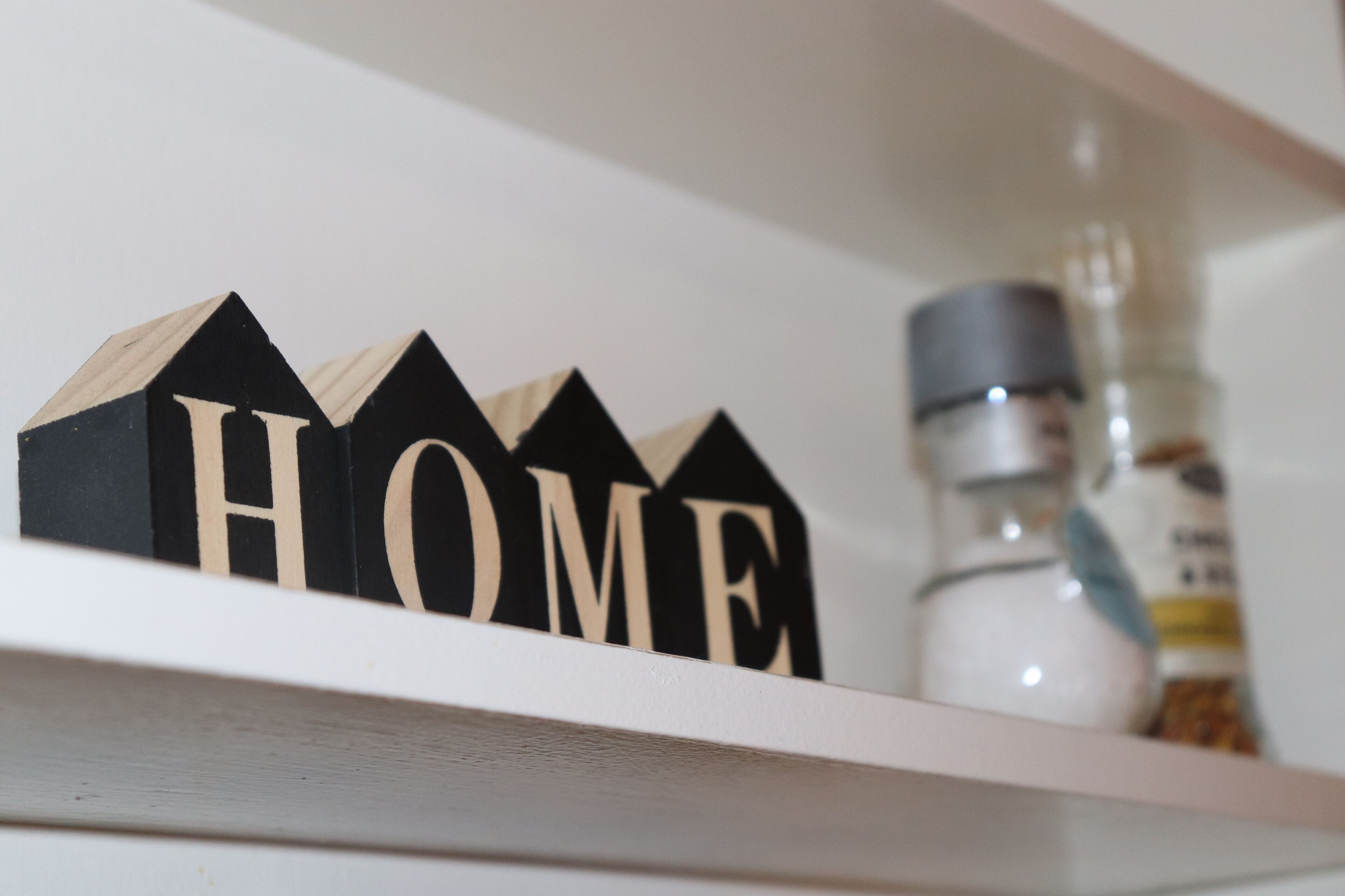 Hyllyllä on puisia, taloa muistuttavia kirjaimia, joista on muodostettu sana ”HOME”, sekä valkoinen pullo, jonka etiketissä on sininen väri.