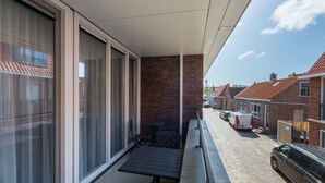 Un balcón con mesa y sillas, un muro de ladrillo y vista a una calle con coches estacionados.