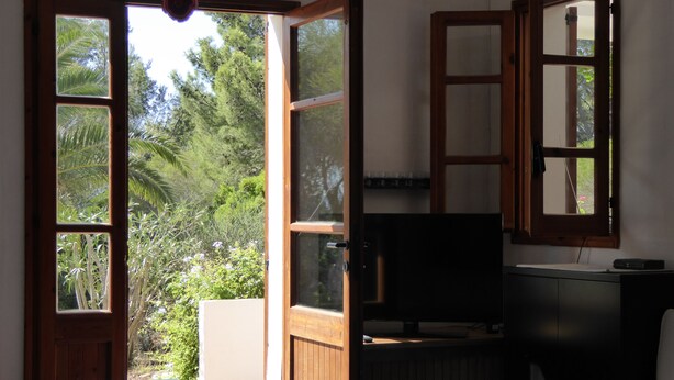 A room with a bed, a TV, and a table with a decorative item. There are open doors leading to a balcony with a view of trees and a garden. - Villa nestled in the countryside with spectacular views for 6 people (Carloforte)