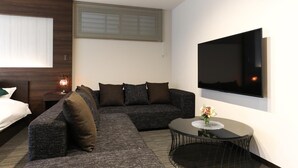 A modern hotel room with a sectional sofa, a small round table with a vase, a flat-screen TV mounted on the wall, and a window with blinds. - GRAND BASE Hakata (Fukuoka)