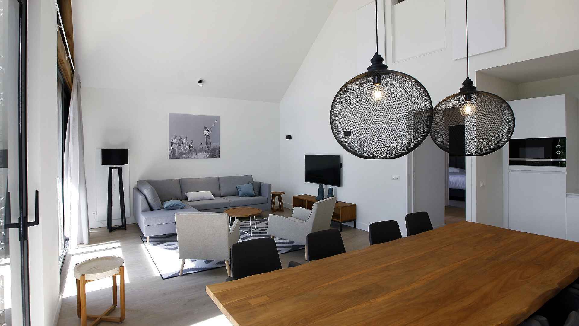 A modern living room with a grey sofa, a wooden dining table, and black chairs. There are two large, round, black pendant lights hanging from the ceiling.