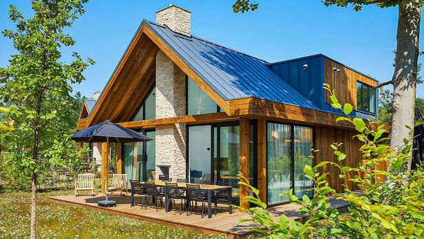 A modern house with a stone chimney, large glass windows, and a wooden deck with outdoor furniture. - Eagle 8p - Four Bedroom Villa, Sleeps 8 (Oosterhout)