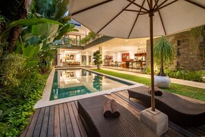 Ein moderner Poolbereich im Freien mit Liegestühlen und einem großen Sonnenschirm, umgeben von üppigem Grün und einem gut beleuchteten Innenbereich.