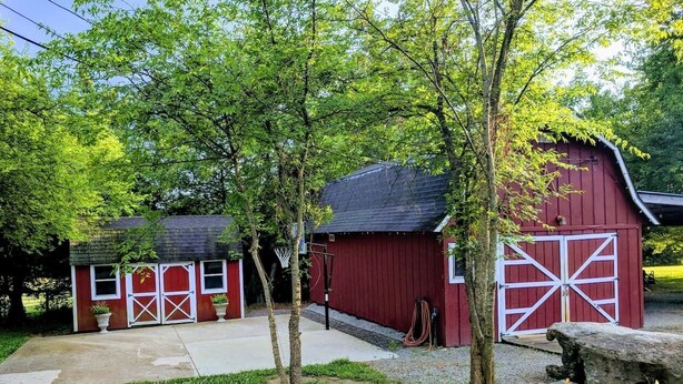 Ein rotes Bauernhaus mit weißen Türen, ein kleinerer roter Schuppen und ein Basketballkorb auf einer grünen Wiese mit Bäumen.
