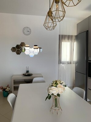 A modern kitchen with a stainless steel refrigerator, a white dining table with a vase of flowers, and a hexagonal mirror on the wall.