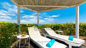 Un espace de détente extérieur abrité, avec deux chaises longues, une petite table sur laquelle se trouve un saladier rempli de fruits, et une vue sur la mer et les collines.