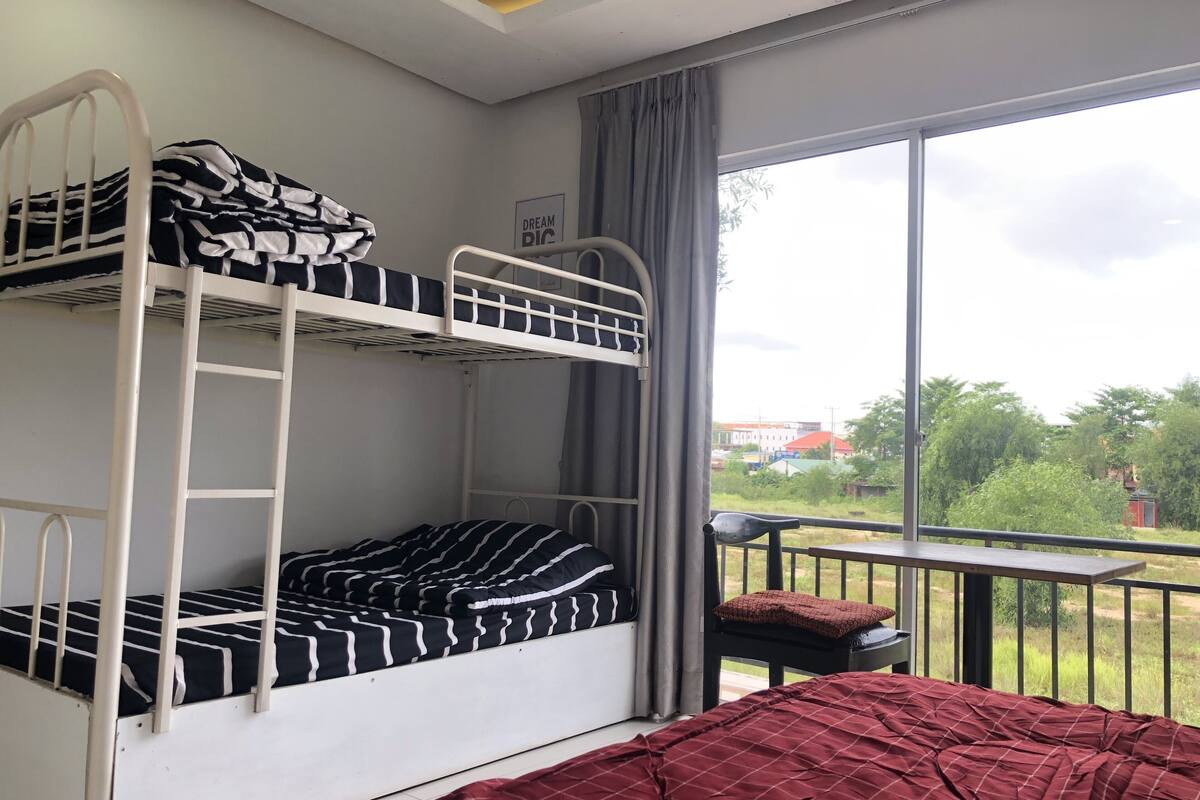 A room with bunk beds, a balcony with a view of greenery and buildings, and a large window with curtains.