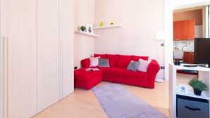 Ein modernes Wohnzimmer mit einem roten Ecksofa, einem kleinen Hocker und einem grauen Teppich. Weiße Regale mit Dekorationselementen und ein Fernseher auf einem weißen Schrank.