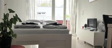 A bedroom with a bed, a desk, a chair, a potted plant, and a window with curtains.