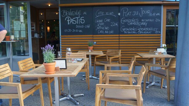 Een terras met houten tafels en stoelen, een krijtbord met de menukaart en een potplant.