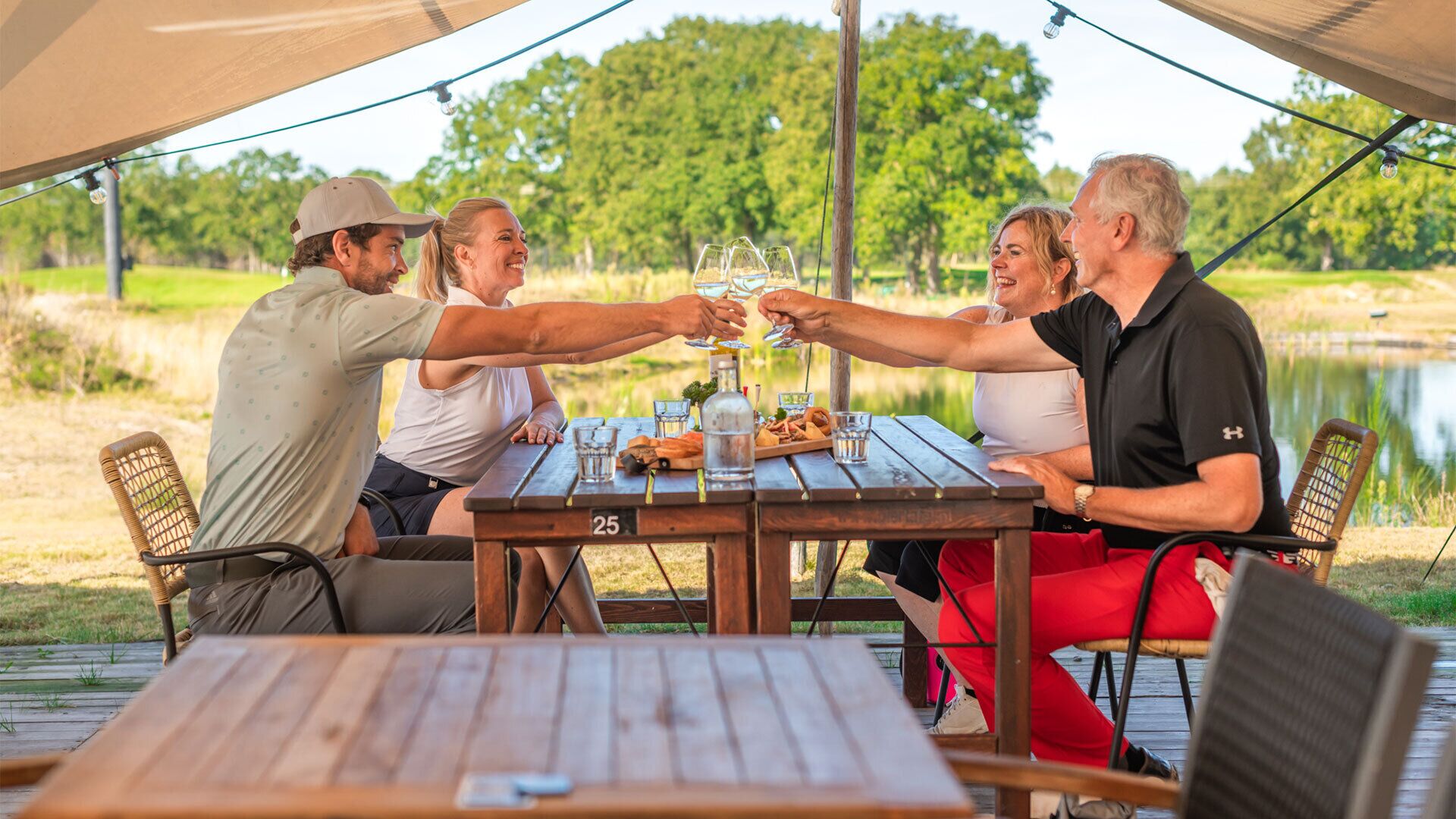 A group of people toasting at a wooden table with a number 25 on it, surrounded by chairs and a picnic setup on a deck with a lake view.