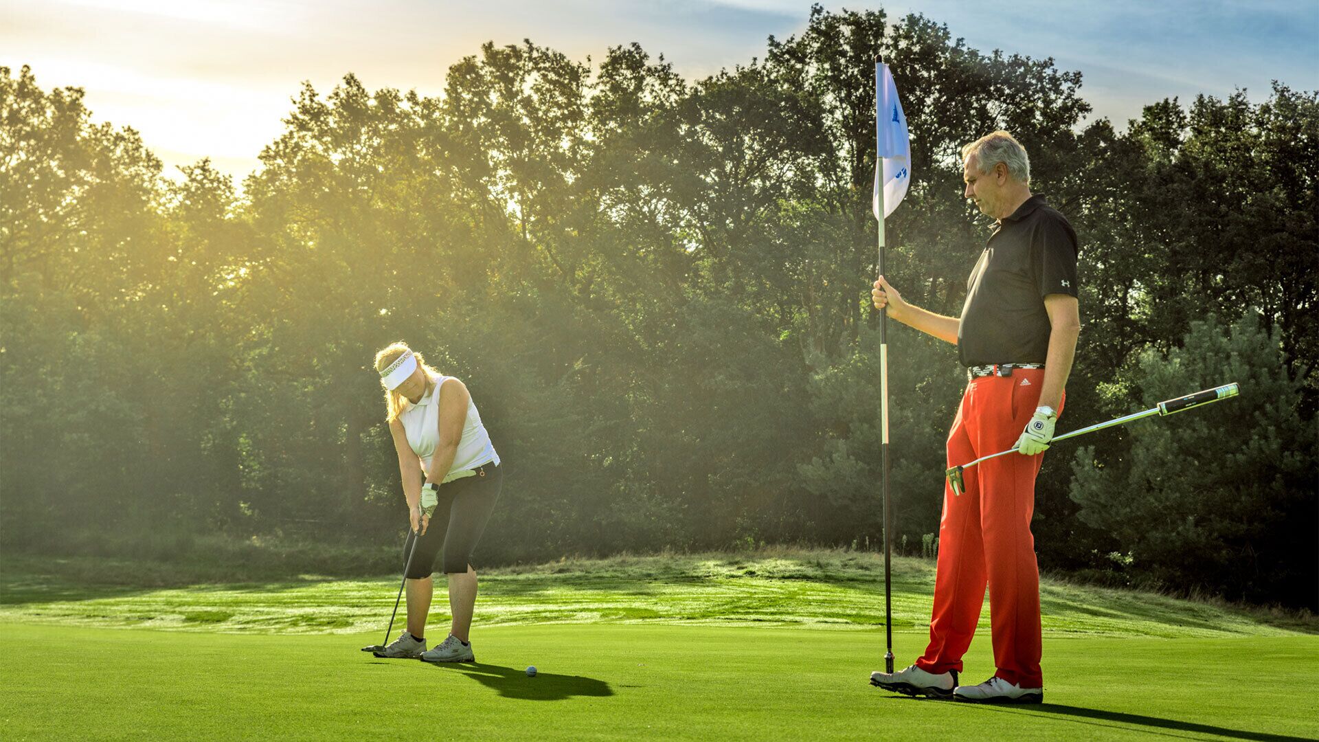 Un campo de golf con un mástil de bandera, un golfista con pantalones rojos y una camiseta blanca, y otro golfista con pantalones negros y una camiseta blanca.