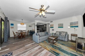 Un salon doté d’un ventilateur de plafond, un coin repas avec une table et des chaises, une cuisine à l’arrière-plan et un tapis aux couleurs vives.