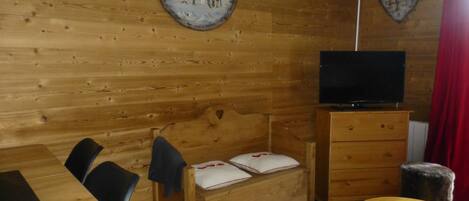 Ein Holzblockhaus-Interieur mit einem hölzernen Bett, einem Fernseher auf einem Standfuß, einem Esstisch und einer Wanduhr.