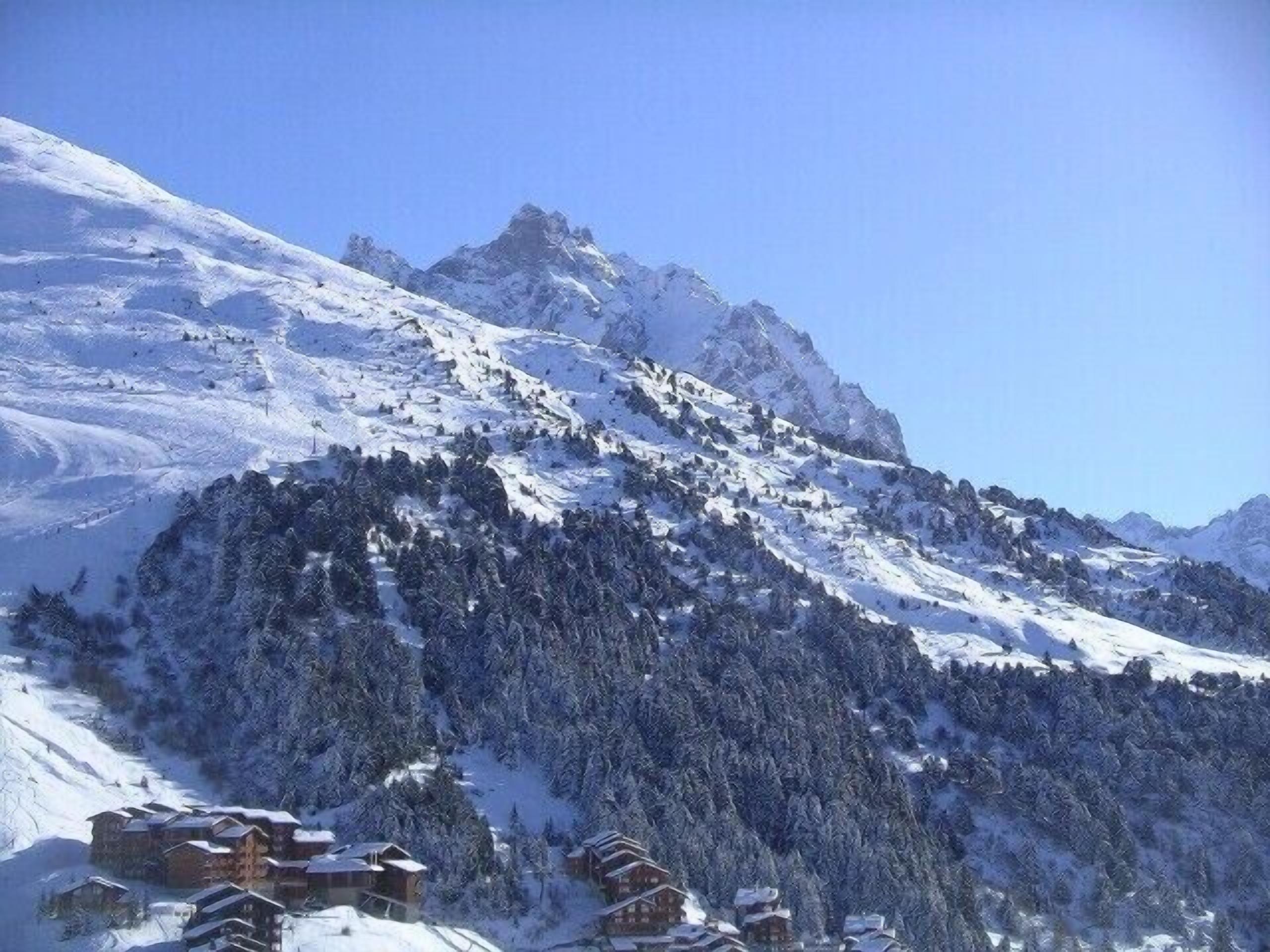 Une montagne enneigée sous un ciel dégagé. On aperçoit des bâtiments au pied de la montagne, probablement un complexe hôtelier ou un chalet.