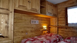 Une chambre de style cabane en bois, avec deux lits, des armoires en bois, un luminaire fixé au mur et une fenêtre avec des rideaux.