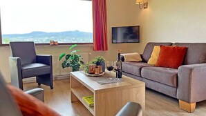 A modern living room with a sofa, armchair, and coffee table. There is a window with a view of the outdoors. - Vulkaneifel Type L-A4 (Gerolstein)