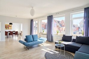A modern living room with a blue sofa, a dark sofa, and a dining area with a table and chairs. - Apartment harbor bay Moba-travel (Travemünde)