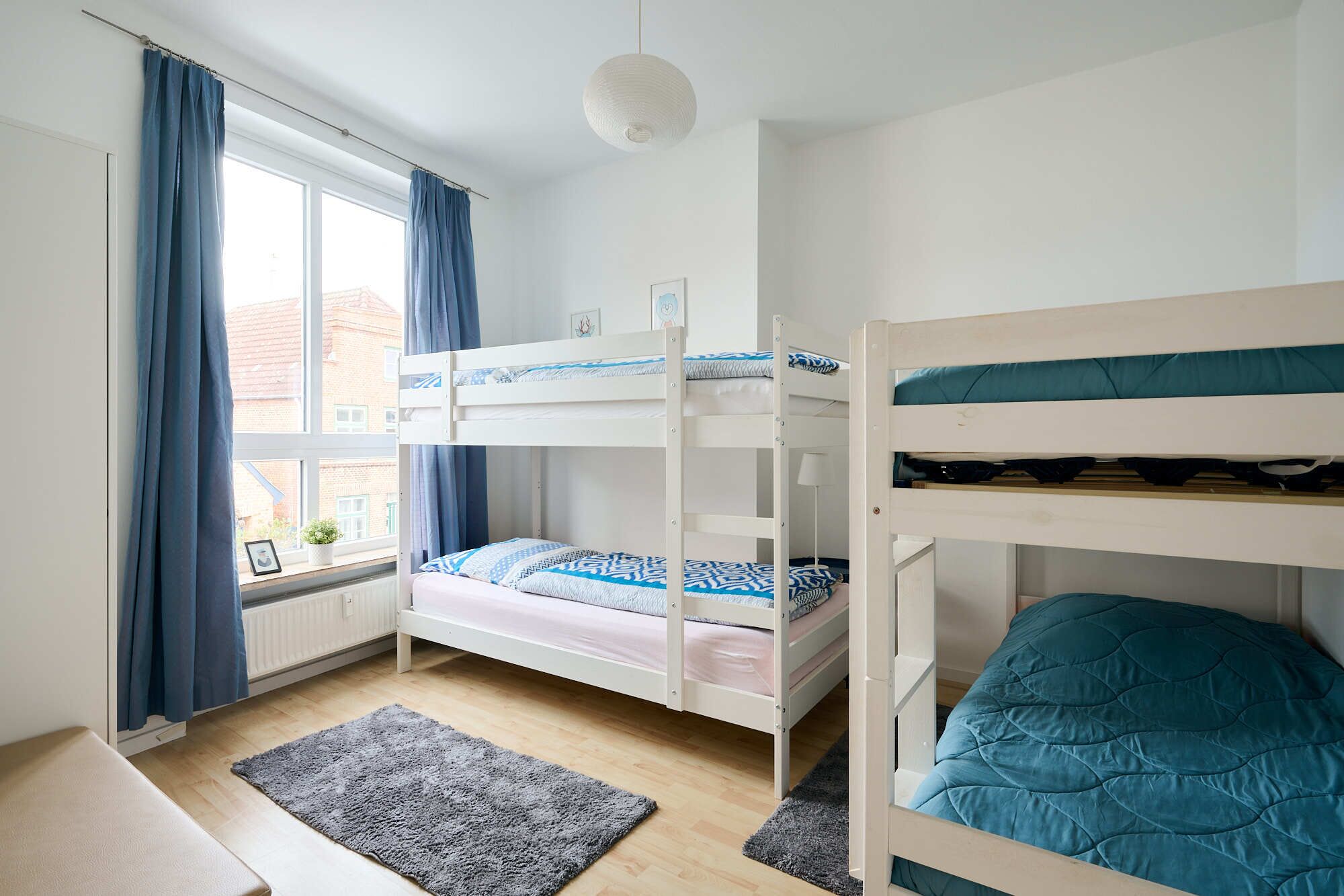 Een slaapkamer met stapelbed, raam, gordijnen, een klein vloerkleed en een witte plafondlamp.