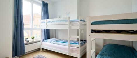 Een slaapkamer met stapelbed, raam, gordijnen, een klein vloerkleed en een witte plafondlamp.