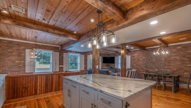 A spacious kitchen with a large island, wooden ceiling beams, and a brick wall. - The Loft on Trade Street | Downtown Tryon | Near TIEC (Tryon)