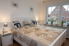 A bedroom with a white bed, a nightstand, a window with a view of a brick house, and a radiator.