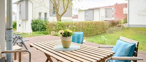 An outdoor patio with a wooden table and chairs, a vase with flowers, and a view of residential buildings in the background.