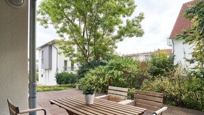 An outdoor patio with a wooden table and chairs, surrounded by greenery and buildings.