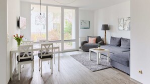 A modern living room with a grey sofa, white dining table, and chairs, a grey rug, and large windows offering a view of the outdoors.