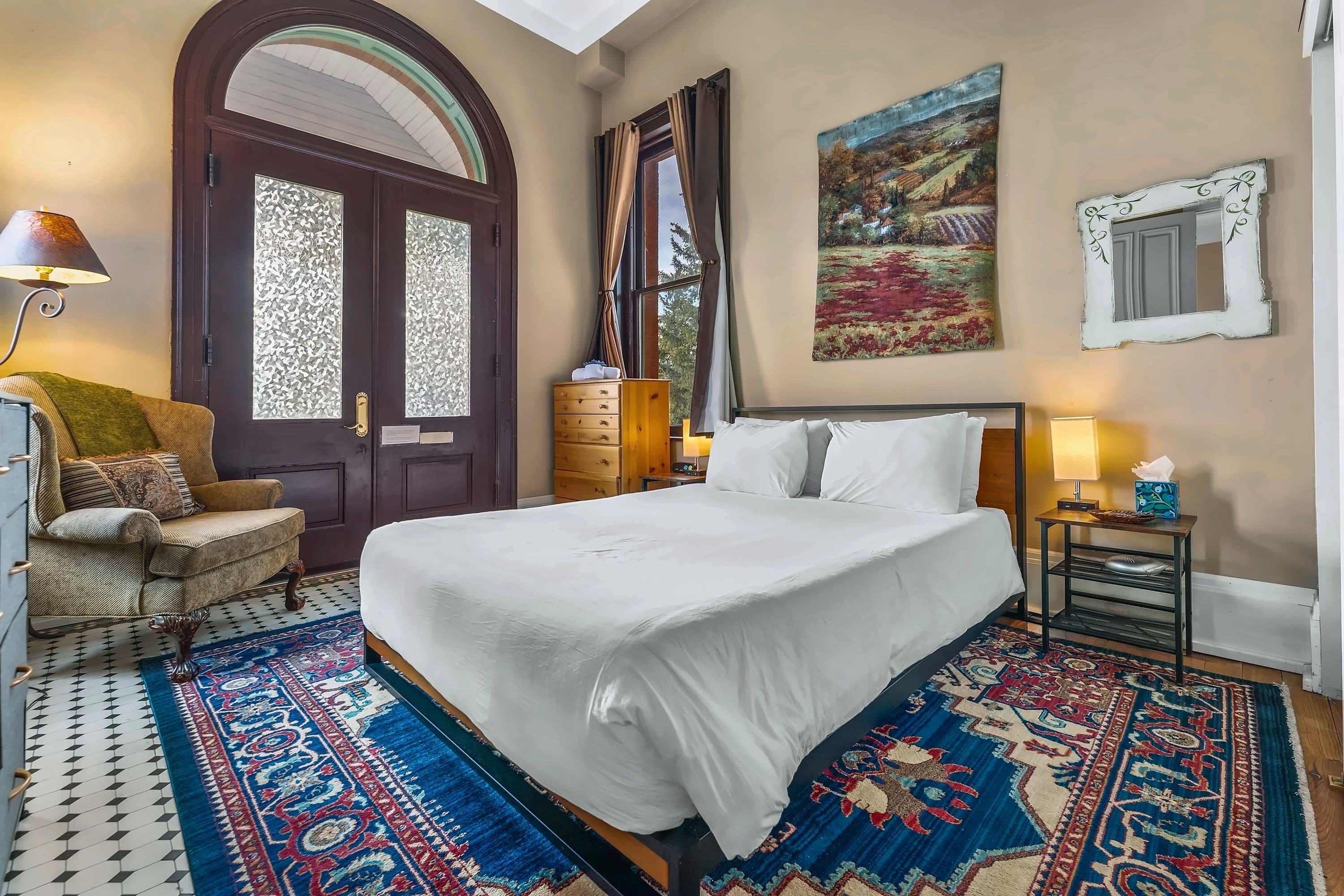 A hotel room with a large bed, a wooden dresser, a chair, a desk with a lamp, and a painting on the wall.