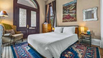 A hotel room with a large bed, a wooden dresser, a chair, a desk with a lamp, and a painting on the wall.