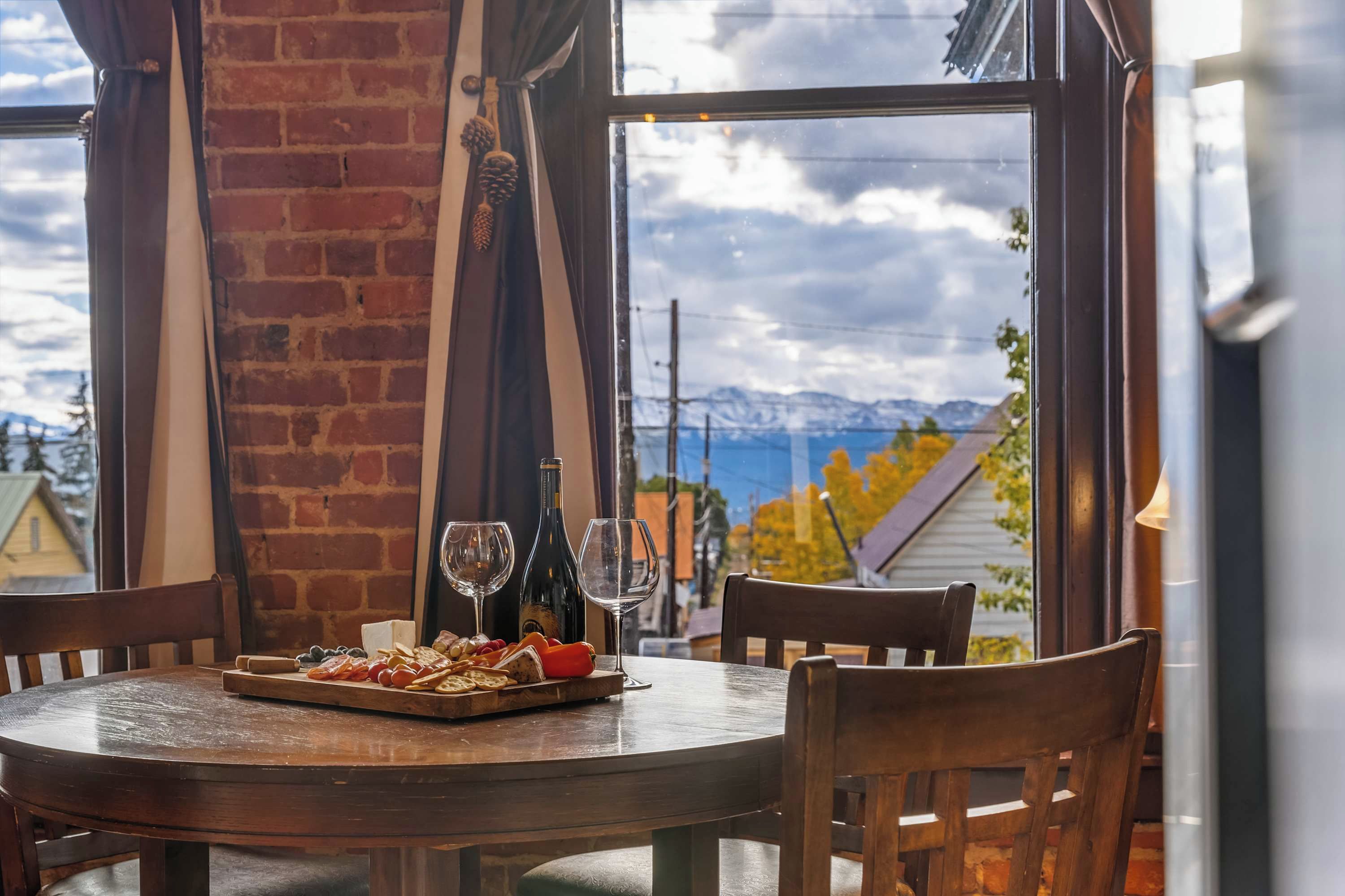 A dining table with a wooden board of snacks and two wine glasses, a bottle, and a candle, set against a brick wall with a view of mountains and a house.