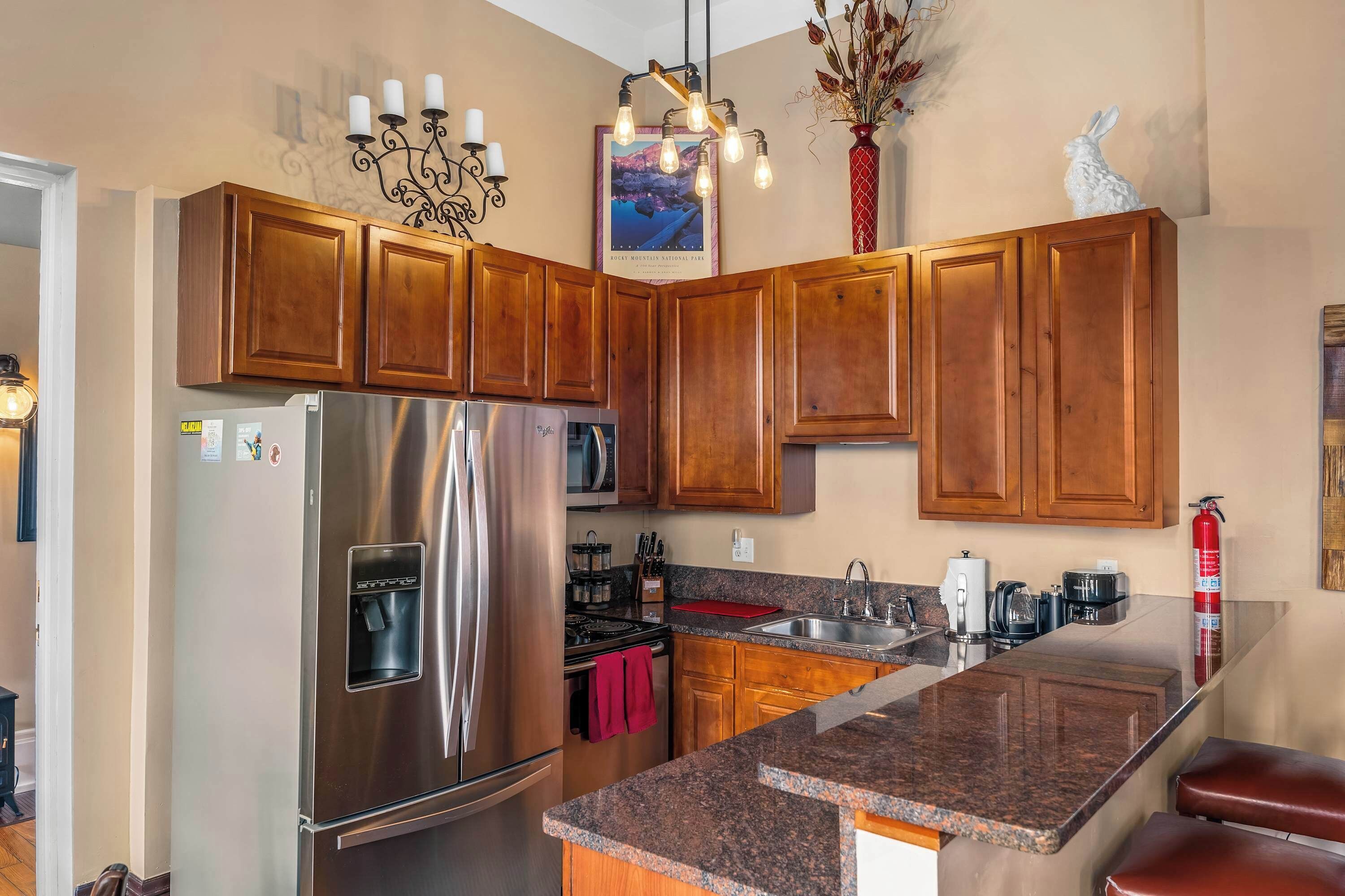 A kitchen with wooden cabinets, a stainless steel refrigerator, a granite countertop, and a hanging light fixture.