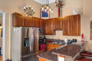 A kitchen with wooden cabinets, a stainless steel refrigerator, a granite countertop, and a hanging light fixture.